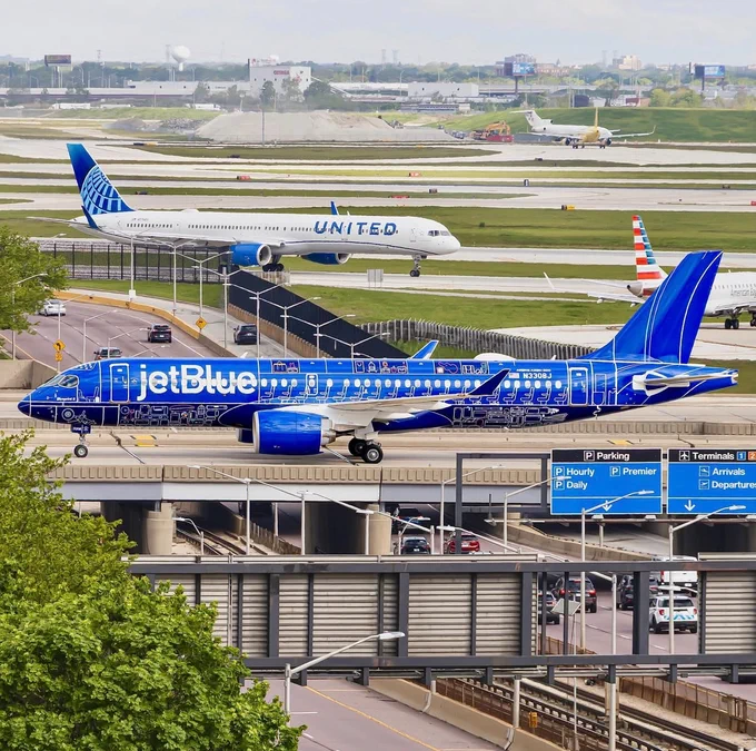 A JetBlue airplane with vibrant blue livery is taxiing on a runway. A United Airlines plane and an American Airlines plane are also visible further back on the tarmac. The airport setting includes roads and directional signs in the foreground, with greenery and other airport infrastructure visible.