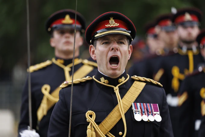 A person in a dress uniform, adorned with multiple medals on the chest, is leading a group of similarly dressed individuals. The person is vocalizing, possibly giving a command, during a ceremonial event. The uniform features gold embellishments and a red and black peaked cap.