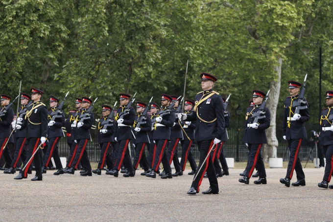 A group of uniformed guards are marching in formation. The guards are wearing dark uniforms with red stripes and caps, carrying rifles over their shoulders. In the background, there are trees.
