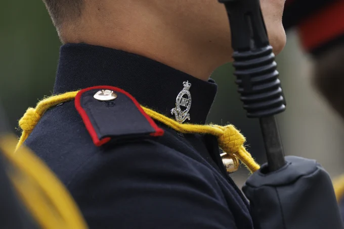 A close-up of a person in a formal uniform features a badge with intricate design details on the collar. The uniform is dark with red and yellow accents.