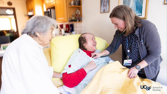 Smiling nurse holds stethoscope to chest of smiling patient lying in bed in their home, while a family member looks on.