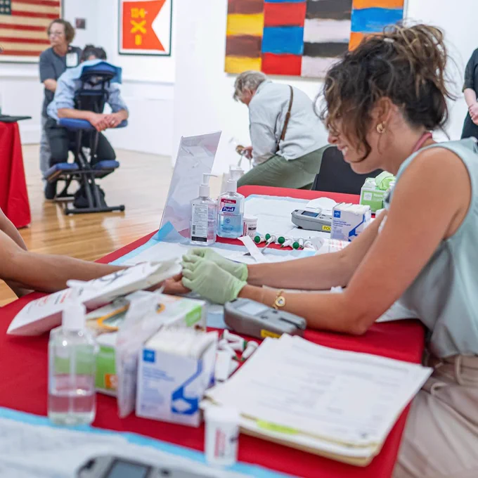An individual is receiving a health check at a table covered with medical supplies, including sanitizers and gloves. There are colorful abstract paintings in the background, along with a visible part of the U.S. flag. Other people are milling around in the art gallery setting.