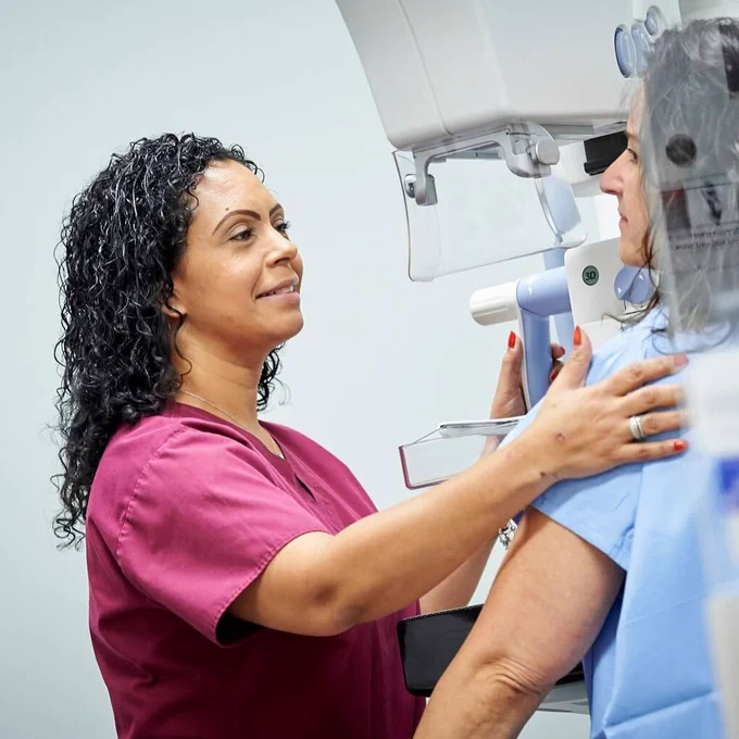 A healthcare professional in maroon scrubs assists a patient with a mammography machine in a medical facility.