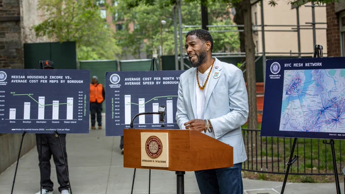 The Public Advocate speaks from a podium, with three posters behind him. 1 shows the median household income versus average cost per mbps by borough. 2 shows average cost per month versus average speed by borough. 3 shows a map of the NYC Mesh network