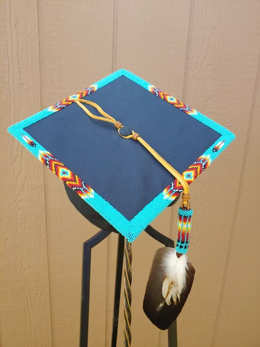 A graduation cap featuring beadwork around the edges and a feather hanging from the cap by a leather strap. 