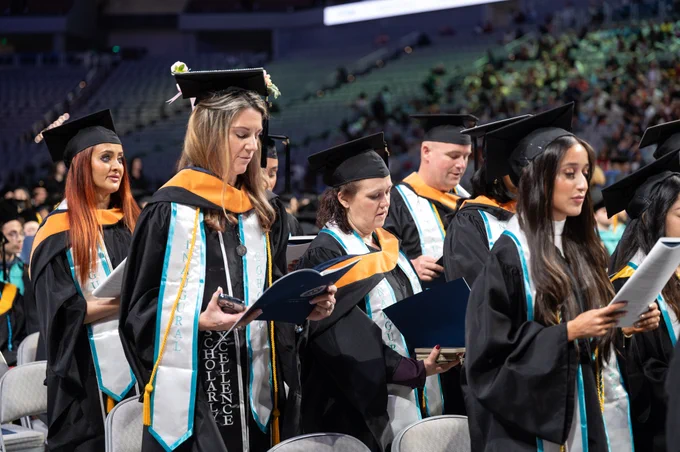 MSN Students in graduation attire, including caps and gowns, is standing in an auditorium. Some are holding programs, and two individuals have sashes with the words "Scholarly Excellence." The seating area is filled with more graduates and attendees.