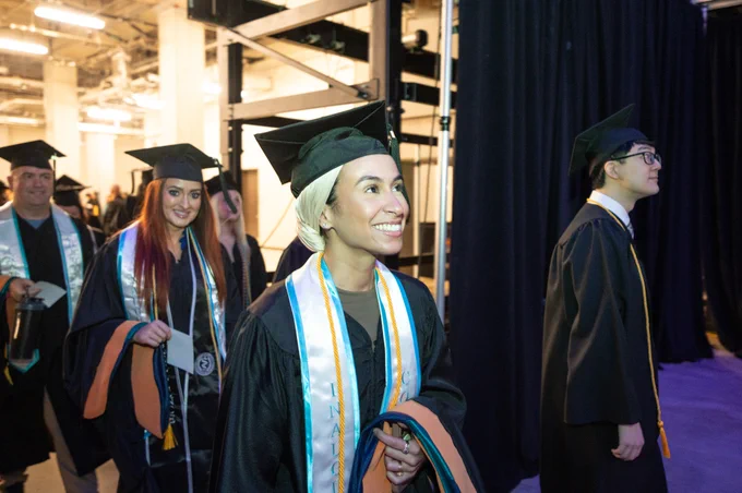 A group of graduates in caps and gowns walk together during a commencement ceremony. They are inside an industrial-style venue with visible lighting fixtures and dark curtains. One graduate in the foreground is smiling and wearing blue and orange honor cords.