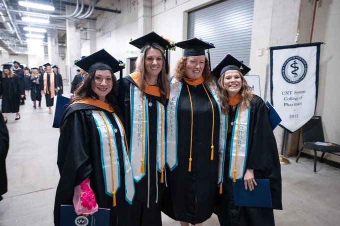 A group of four graduates in academic regalia smiles for a photo indoors. They are wearing black caps and gowns with white stoles featuring the word "Doctoral." A banner in the background reads "UNT System College of Pharmacy 2013." Other graduates can be seen in the hallway.