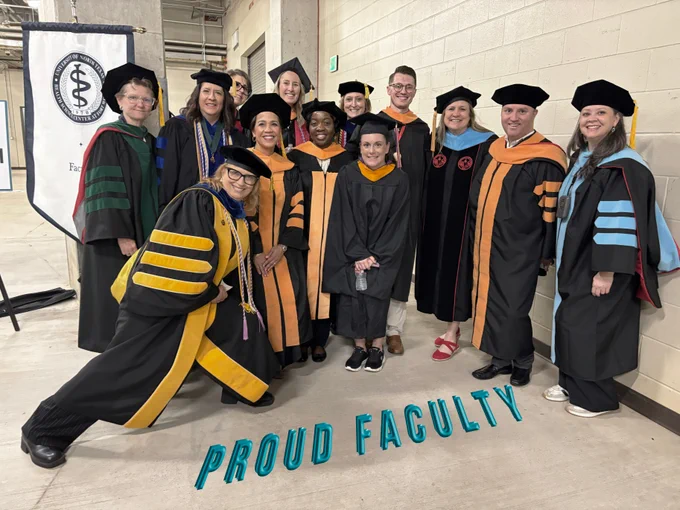 A group of faculty members in academic regalia pose together. They are standing indoors by a wall with a banner featuring a medical symbol and the word "Faculty." Text on the floor reads "Proud Faculty.