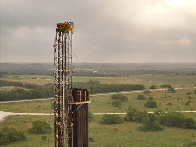 A tall oil drilling rig stands in a green landscape with scattered trees under a cloudy sky, indicating a rural setting. 