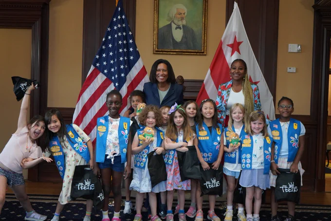 Mayor Bowser and DPR Director Thennie Freeman stand in a posed smiling shot with a dozen Girl Scouts in front of the US and DC flag.