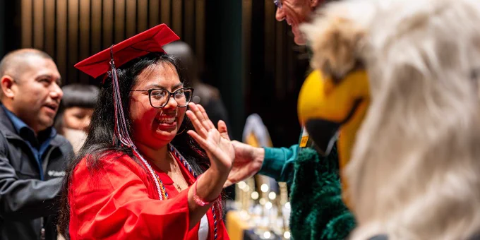A person in a red graduation cap and gown smiles and high-fives a costumed mascot that resembles an eagle.