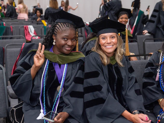 Two individuals in graduation attire smile for the camera, with one holding up a peace sign. They are seated among a group of graduates wearing black gowns and caps.