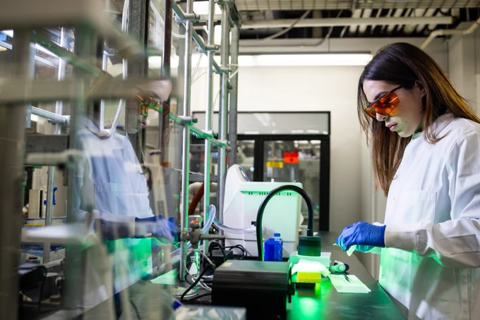 A student works in a lab, measuring liquids while wearing a white coat, safety glasses and gloves.