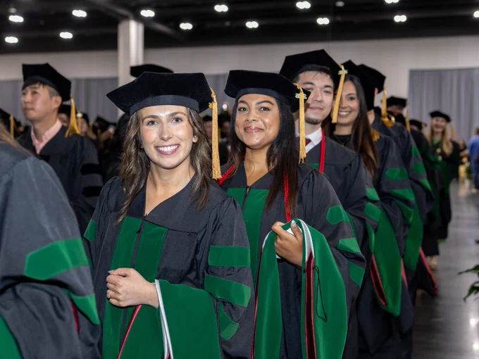Graduates in academic regalia standing in a line at a ceremony, with some wearing green and black gowns and mortarboards.