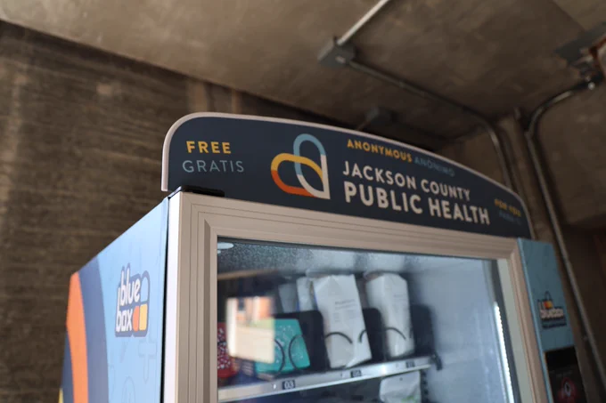 A vending machine labeled "Jackson County Public Health" and "Bluebox." It contains health-related products with signage indicating free availability in both English and Spanish.