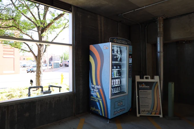 A vending machine labeled "Bluebox" is situated in a parking garage. It offers health products and is positioned next to a sign with information about public health services. Sunlight filters through a large window, revealing an outdoor view with a tree and building.