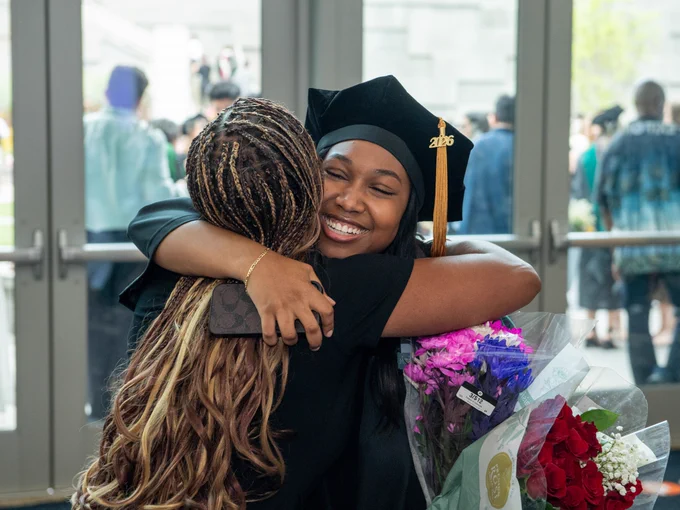 A graduate in a cap and gown joyfully hugs another person while holding a bouquet of flowers. The setting appears to be indoors, possibly a graduation venue with people in the background.