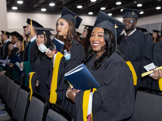 A group of graduates in caps and gowns stand in rows at a ceremony. They are holding diplomas and wearing proud expressions, with colorful cords draped around their necks. The background shows a large indoor auditorium filled with rows of seating.