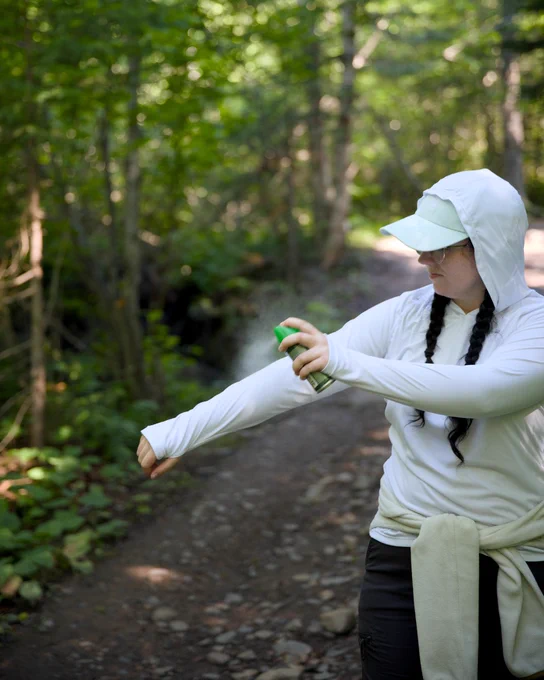 Woman wearing a white long sleeve top is putting on bug spray while standing on a trail in the forest.