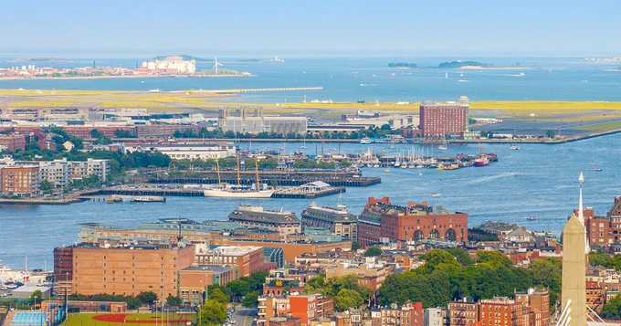 A scenic view of Boston Harbor, featuring the USS Constitution docked at a pier. The image includes buildings along the waterfront, greenery, and distant islands with the expansive ocean in the background.