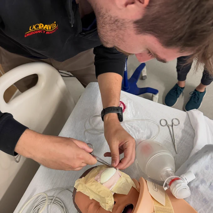A resident in his UC Davis Emergency Medicine jacket practices lateral canthotomy on a mannequin with a simulated eyeball created using a hard-boiled egg.