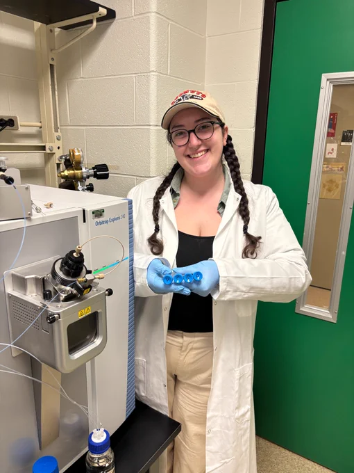 Grace Semaan works smiles at the camera. She is in a lab and is holding samples and wearing a lab coat. 