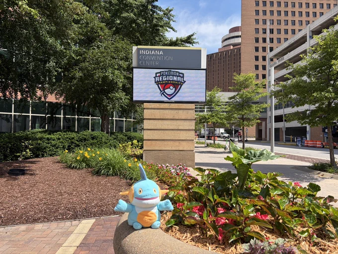 A small marshtomp plush sits on a stone ledge outside the Indiana Convention Center in Indianapolis. In the background, a large digital sign displays the Pokémon Regional Championships logo, with the Hyatt Regency hotel and surrounding city buildings visible on a sunny day.