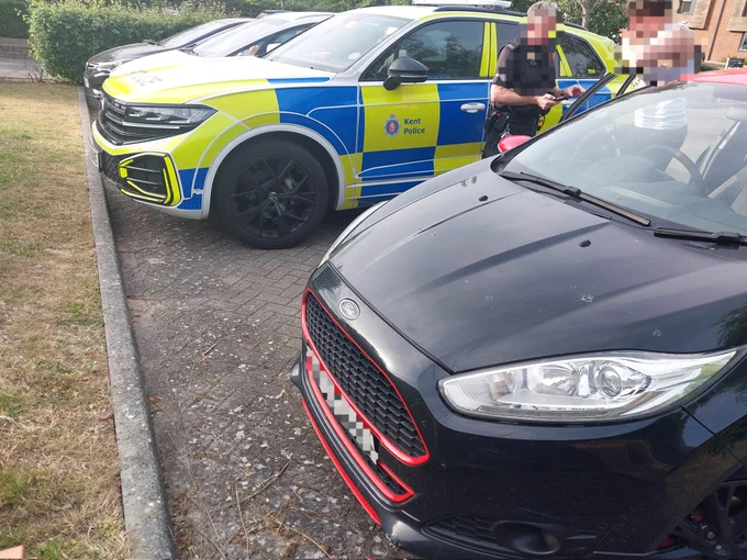 Picture of a Black/Red Ford Fiesta with an officer speaking to the driver reporting him for the offence of no insurance. Behind is TD16 a marked Volkswagen Touraeg