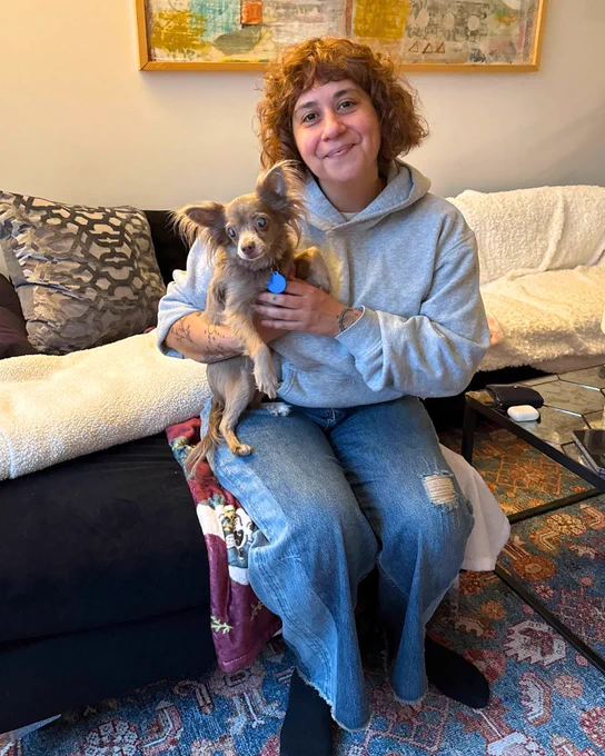 a person holds Gracie on their lap. both are inside on a black and white couch. the person is smiling.