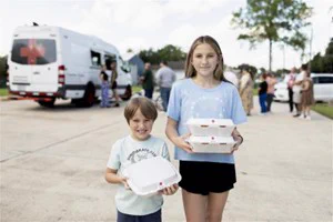 Children standing outside hold stacked food containers, with a Red Cross vehicle and several people in the background.