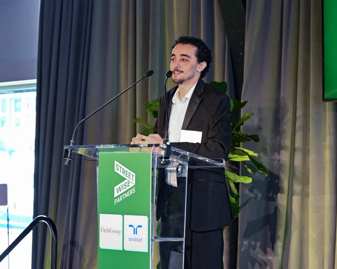 Person in a black suit speaking at a podium with Street Wise Partners branding during an indoor event.