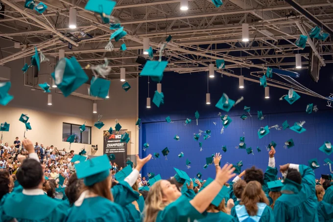 Graduates in teal robes tossing caps into the air in an indoor ceremony setting with decorations and lighting.