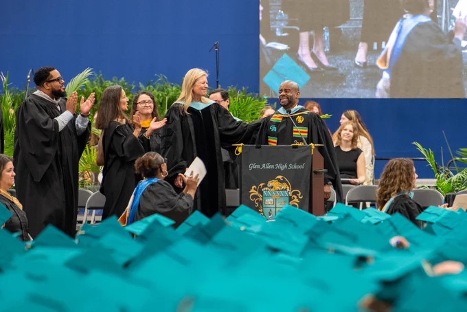 A graduation ceremony with faculty and students in graduation gowns and caps. They stand on a stage surrounded by a blue backdrop and greenery.