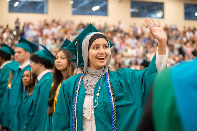 A smiling graduate wearing a green graduation gown and cap raises a fist in celebration at a crowded graduation ceremony.