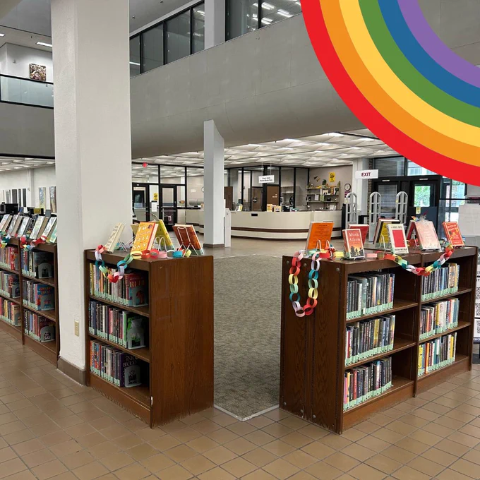 The Sims Library bestseller bookshelves on the first floor. On top of the bookshelves, books are displayed with colorful covers to form a rainbow. Rainbow colored paper chains are also draped across the tops of the bookshelves. The front circulation desk and front doors of the library are visible in the background. 