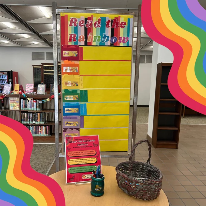 A yellow poster in Sims Library styled to look like a bookshelf. It is titled "Read the Rainbow" and has eight shelves, each assigned a color: red, orange, yellow, green, blue, purple, pink, and black. A few people have added colorful slips of paper to the matching shelf. On the slips are the titles of books that people read with that color cover. A small table in front of the poster has pens, a basket, and instructions for adding a book to the poster. The Sims Library bestseller bookshelves are visible in the background. 