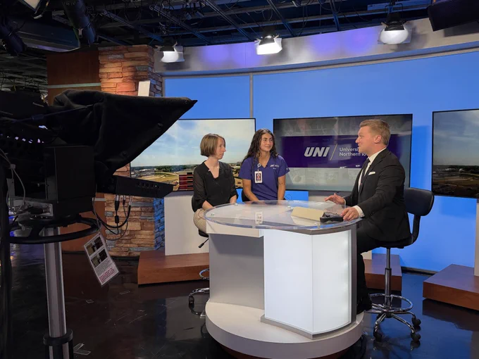 Three people sit at an interview desk inside a TV studio.