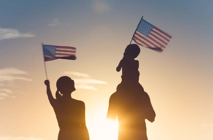 Two people and a child hold American flags silhouetted against a sunset sky. The child sits on one person's shoulders, with a flag waving in the wind.