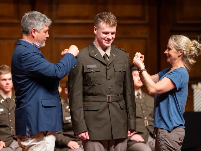 A person in a military uniform stands smiling as two individuals attach insignia to their shoulders. They are indoors, and several people in similar uniforms are seated in the background.