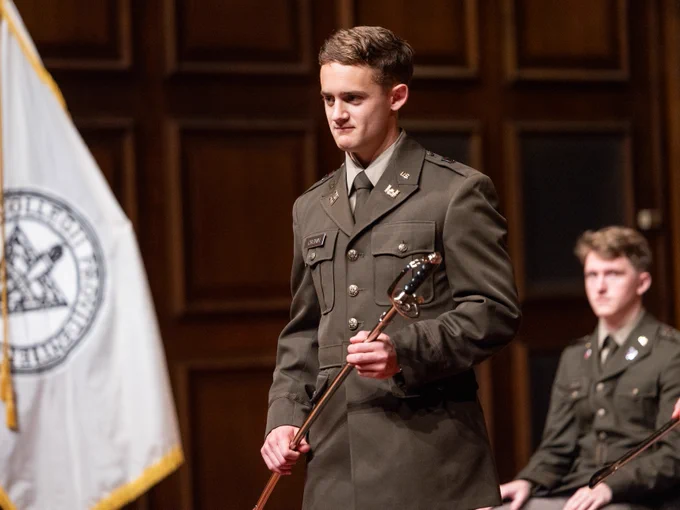 A person in a military uniform holds a ceremonial sword, standing on a stage. Another person in uniform sits behind them. A flag with a seal featuring crossed swords is in the background.