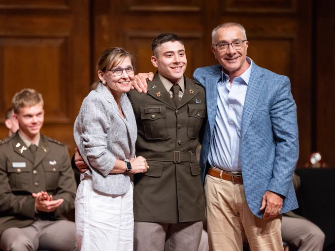 Three people stand posing and smiling for a photo on stage at a ROTC commissioning event. The man in the middle of the two parents is dressed in military attire.