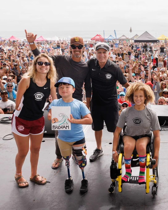 Group on stage at a beach event, including a child with prosthetic legs and a person in a wheelchair, with a cheering crowd behind.