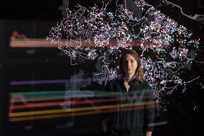 A person stands in front of an illuminated map with various colored points. In the foreground, multi-colored line graphs are visible. 