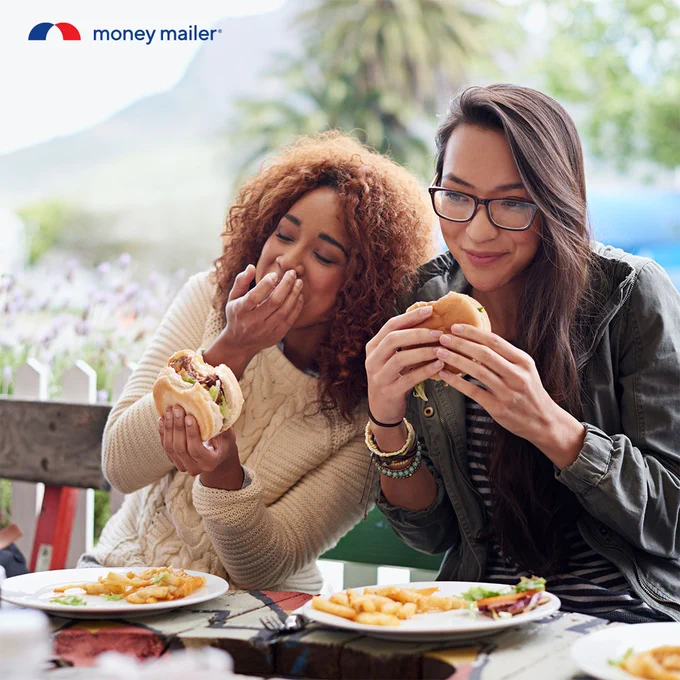 Two friends enjoying burgers and fries outdoors at a wooden table with a scenic background.