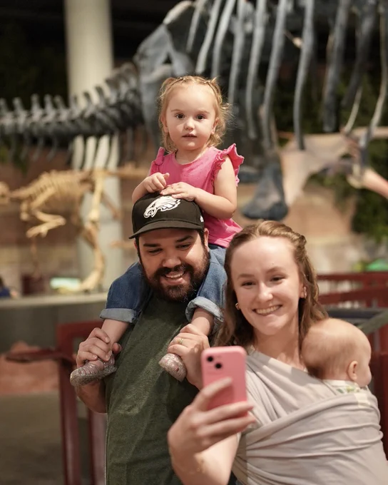 Family of four taking a selfie inside a museum with dinosaur skeletons displayed in the background.