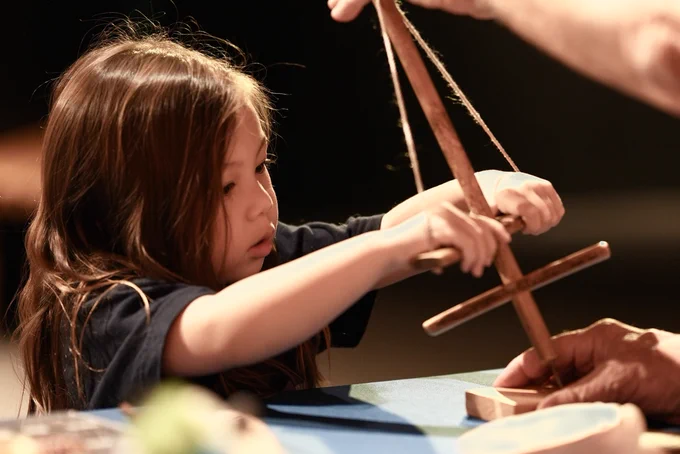 A child carefully holds a wooden drill while an adult guides their hands during a hands-on activity about prehistoric tools. 