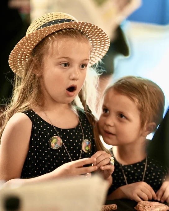 Two young children wearing matching black polka dot dresses and necklaces, one with a straw hat, standing close together indoors. The older girl has a look of wonder on her face.