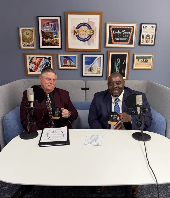 Two men in suits sit on a blue couch behind a white table with microphones, holding mugs in a podcast studio decorated with framed city-themed artwork.