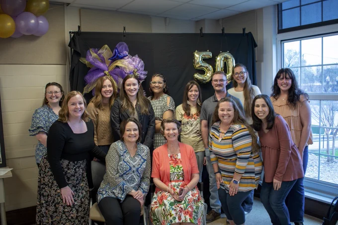 A group of people are gathered in a room decorated for a celebration. There are gold balloons forming the number "50" and purple ribbons in the background. Two individuals are seated in front, while others stand behind them, all smiling.